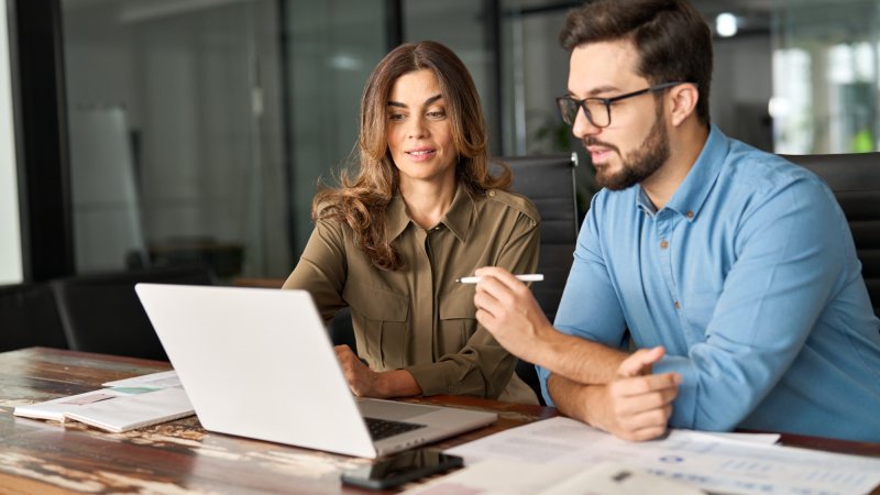 Two colleagues collaborate over a laptop in an office, a woman explaining something to a younger colleague as they review information together