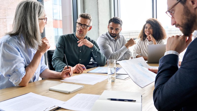 A diverse group of international executives gathered around a conference table, actively reviewing documents and discussing strategy during a collaborative business meeting.