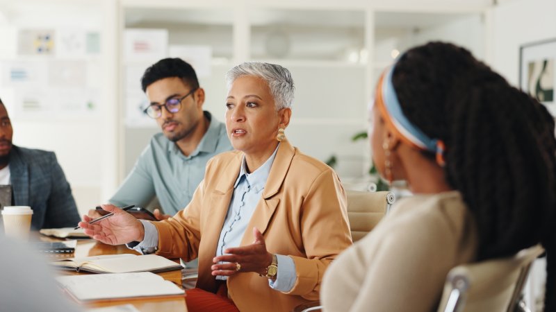 A senior government affairs director with gray hair in an orange blazer leads a strategy discussion with her diverse team around a conference table